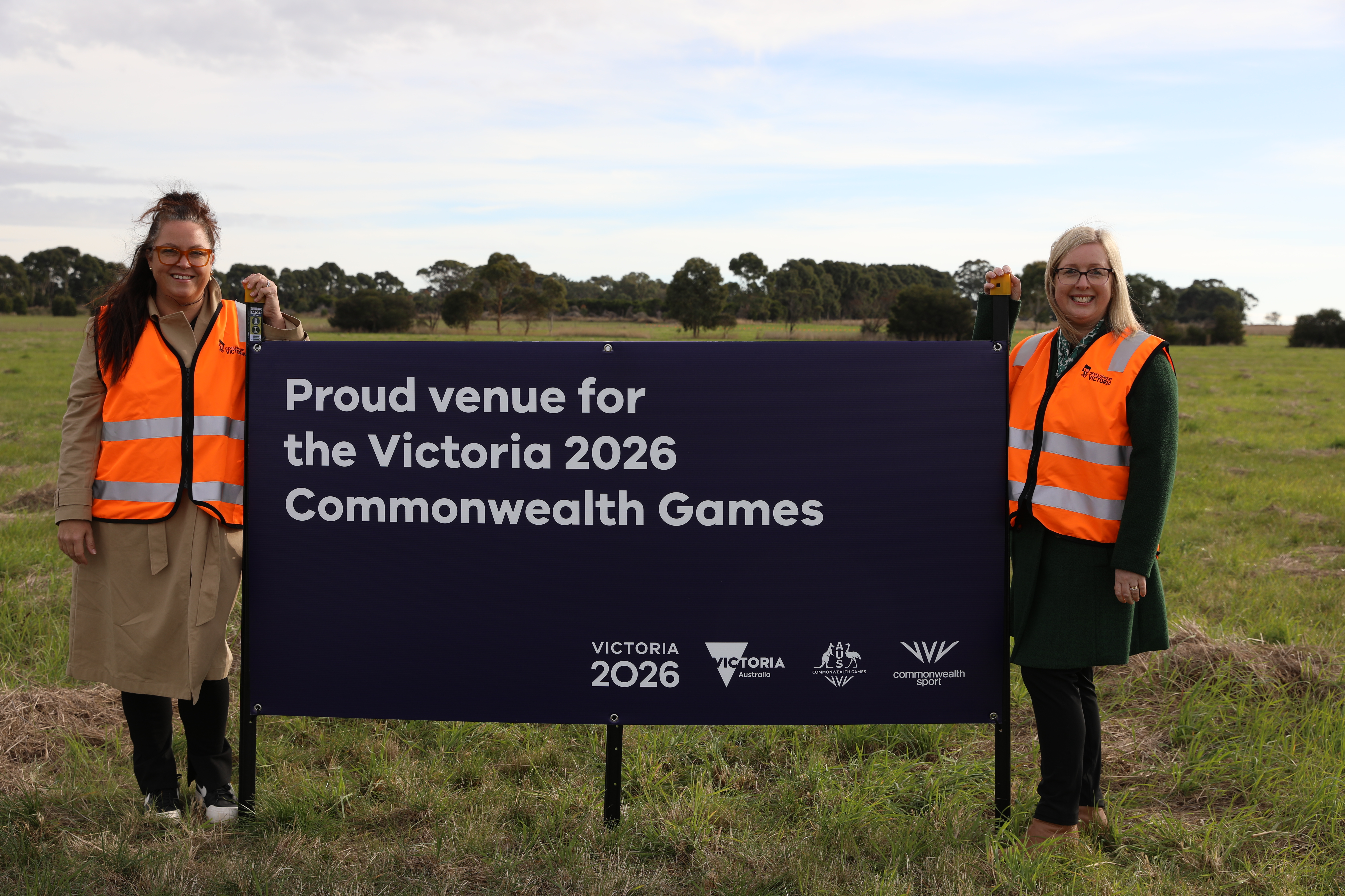 Two white women smiling to camera standing with a sign that says Proud venue for the Victoria 2026 Commonweath Games.