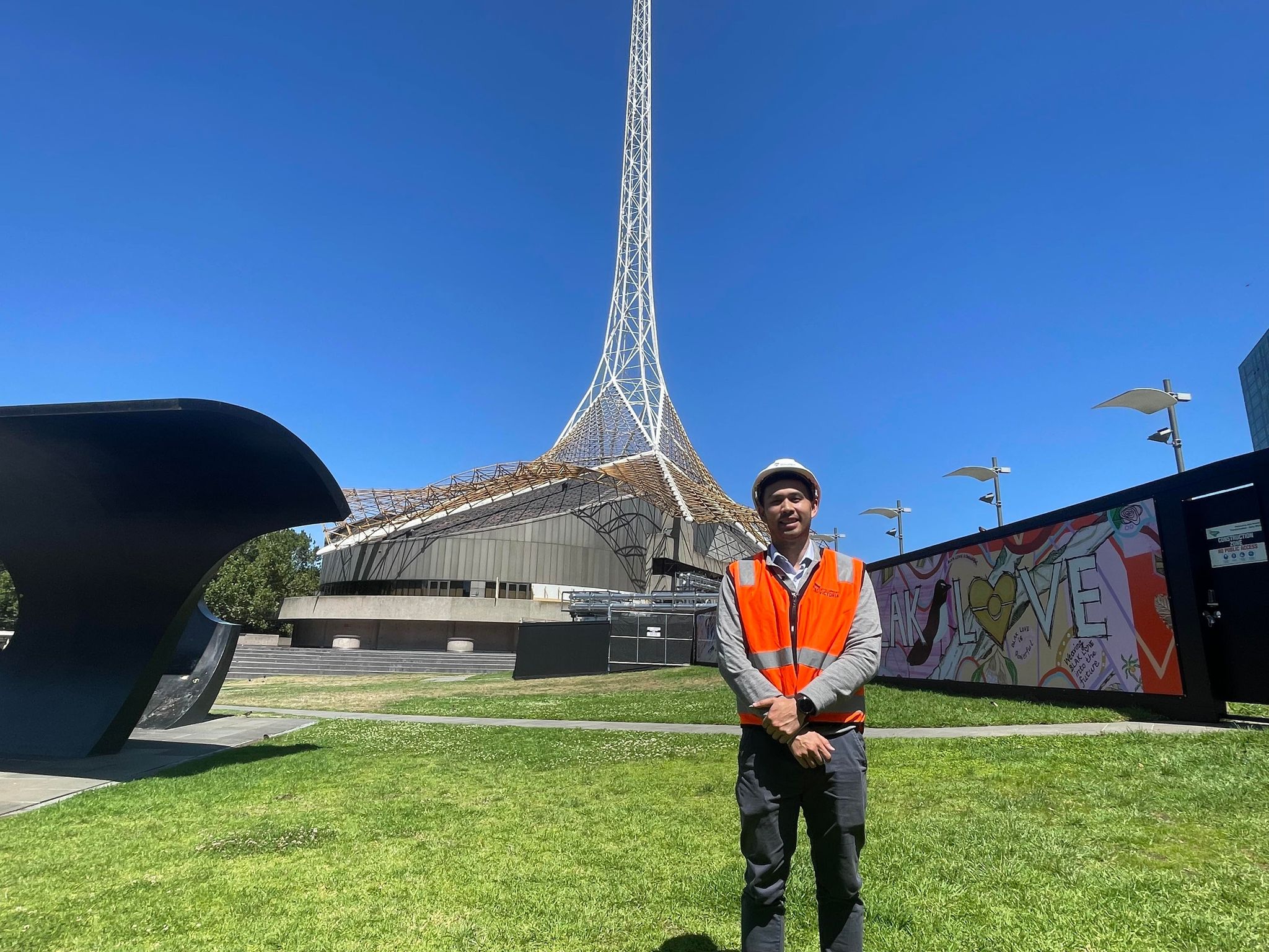 A man wearing orange high vis and a hard hat smiling at camera in front of the Melbourne Arts Precinct spire