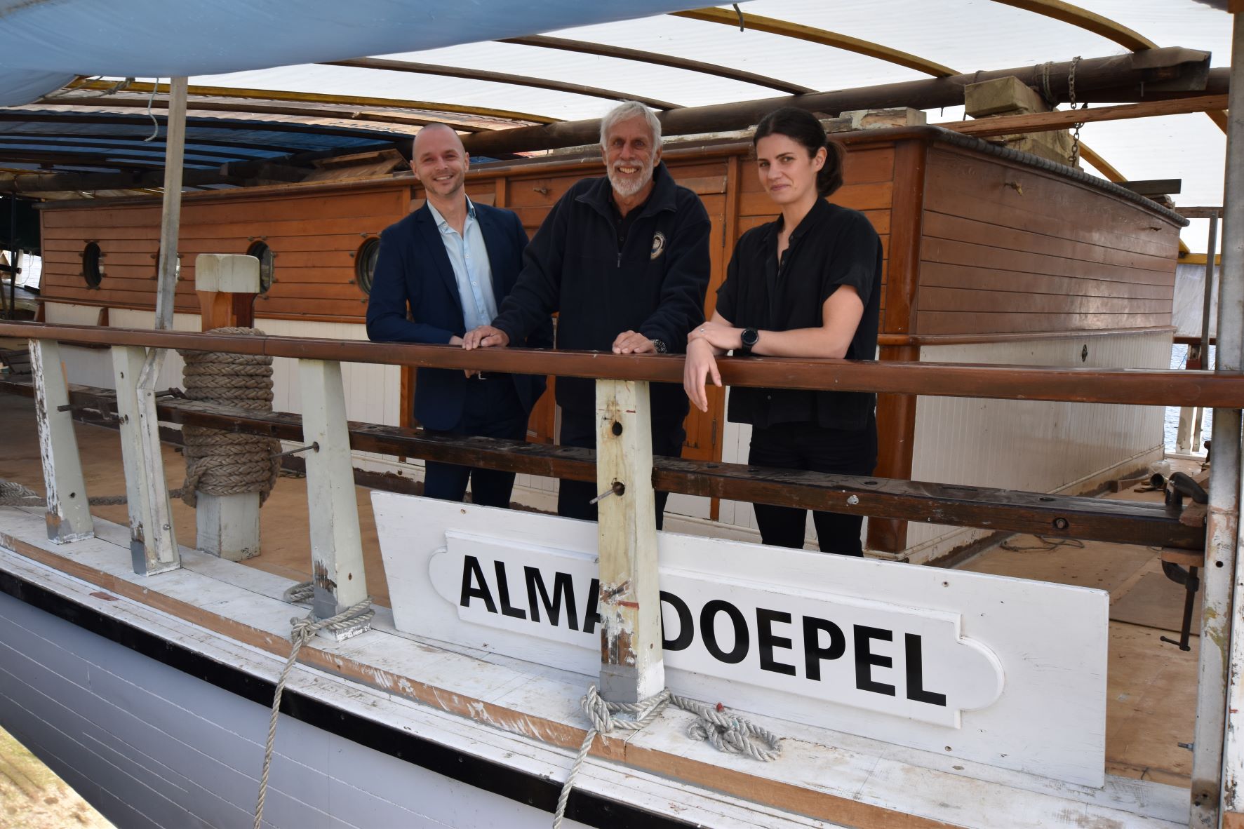 Two men and one woman standing on a boat, hands on the railing and an Alma Doepel sign in front of them
