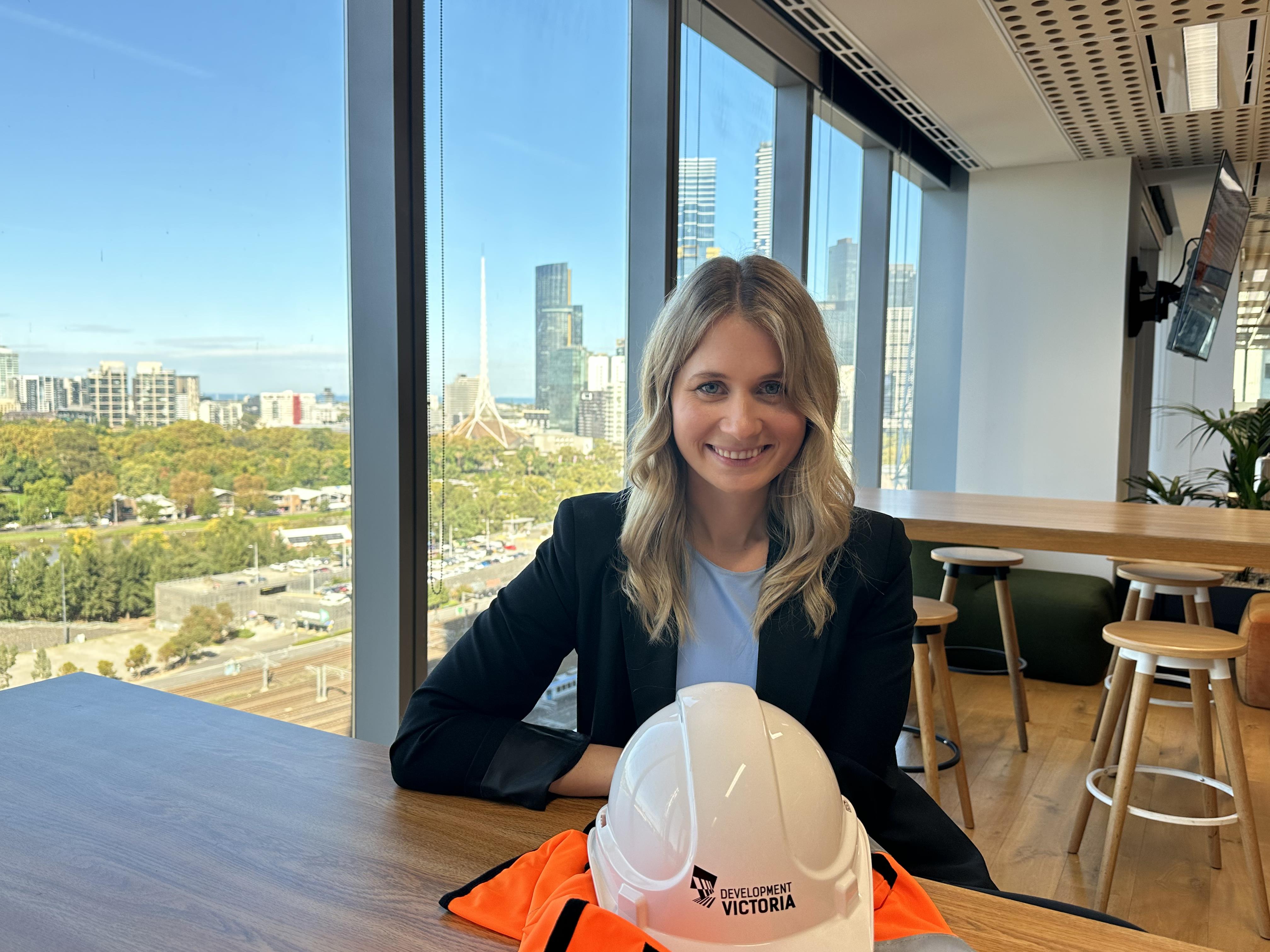A white female with blonde hair smiling to the camera with a white hard hat with Development Victoria written on it in front of her