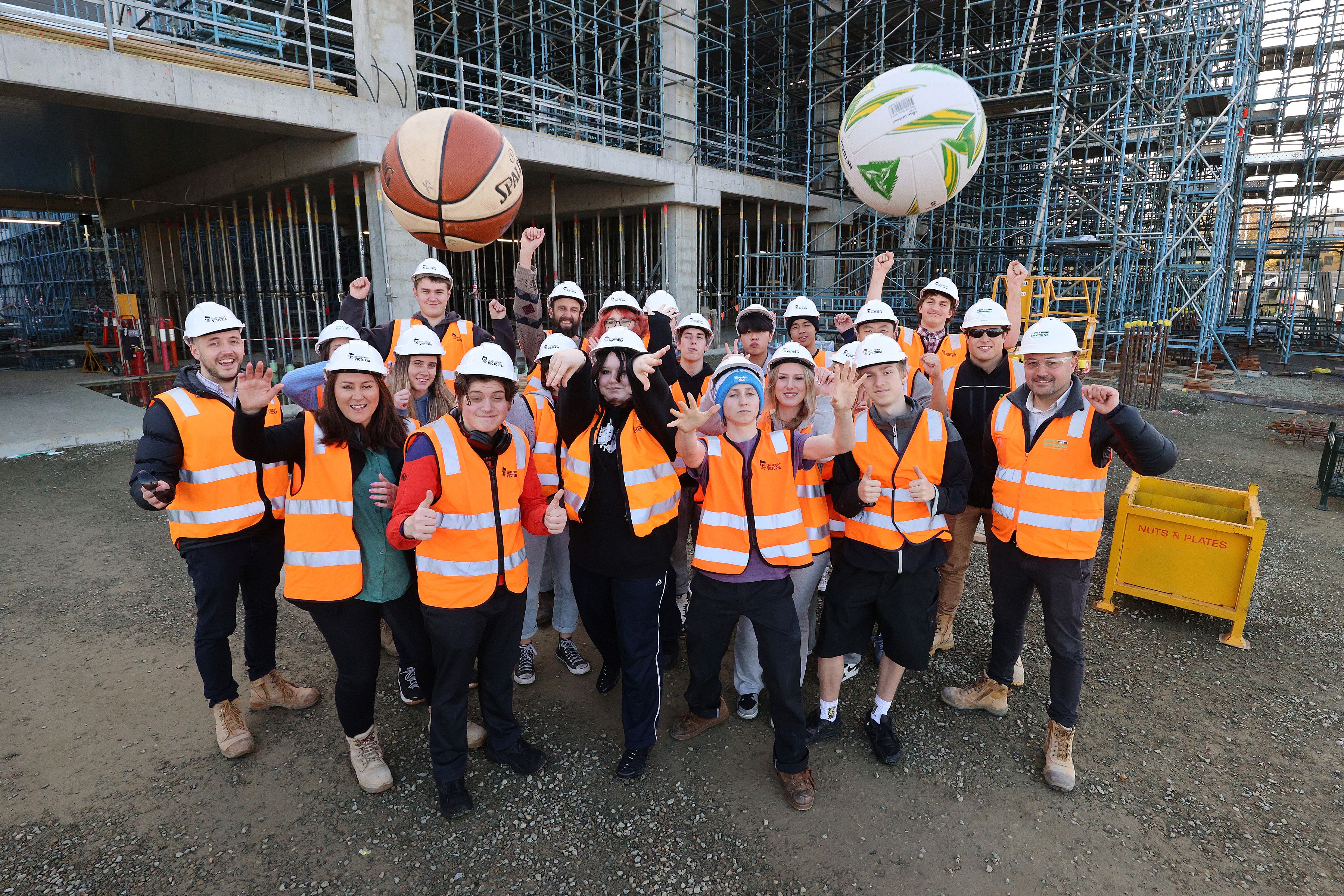 A group of high school students wearing high vis and Development Victoria helmets standing in front of a construction site throwing a basketball and volleyball